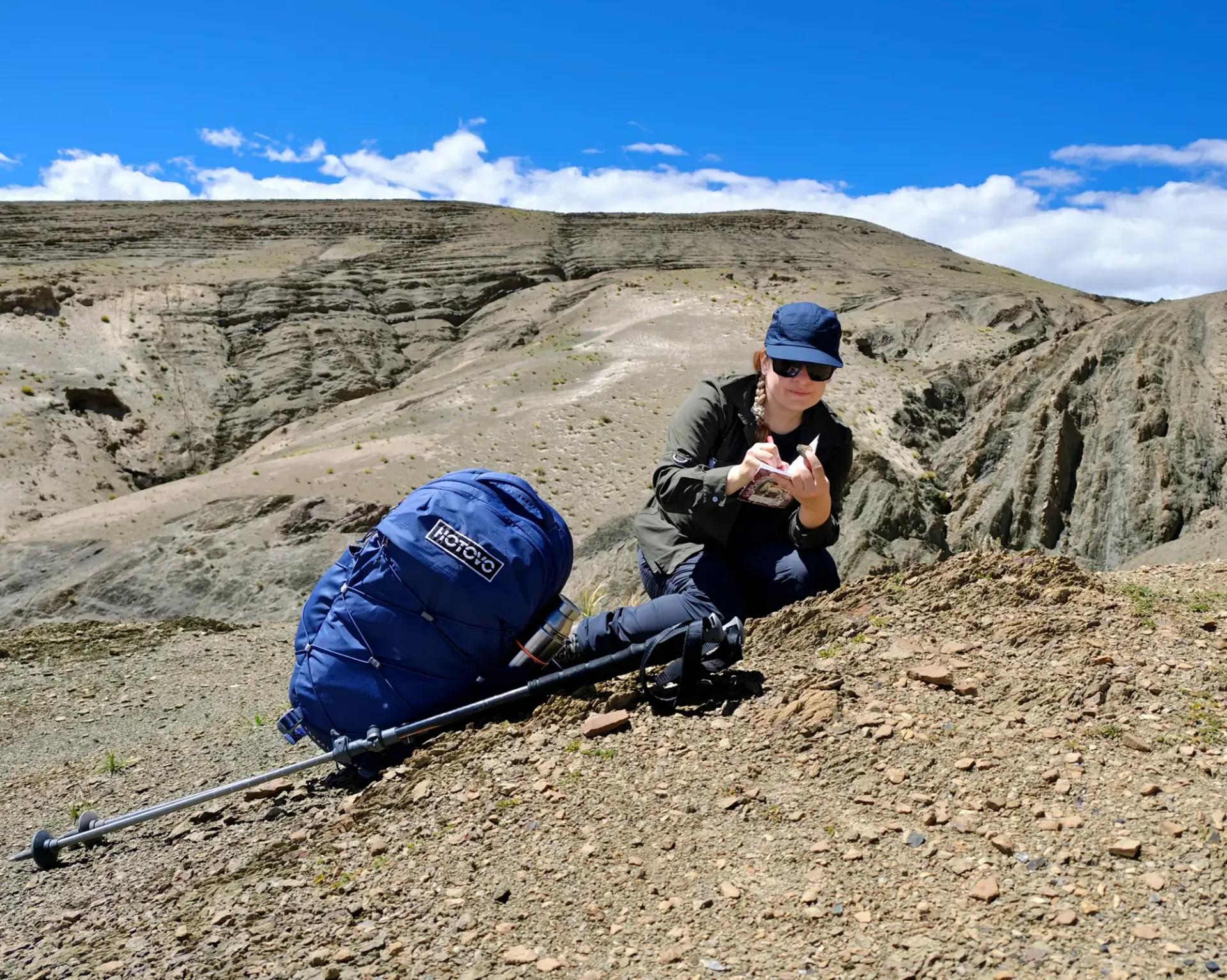 Dr. Michaela Musilova on her Astro Seven Summits mission. Doing a reaearch. Besides her is hoking backback with Hotovo logo. She is squatting on the hill and rocks.
